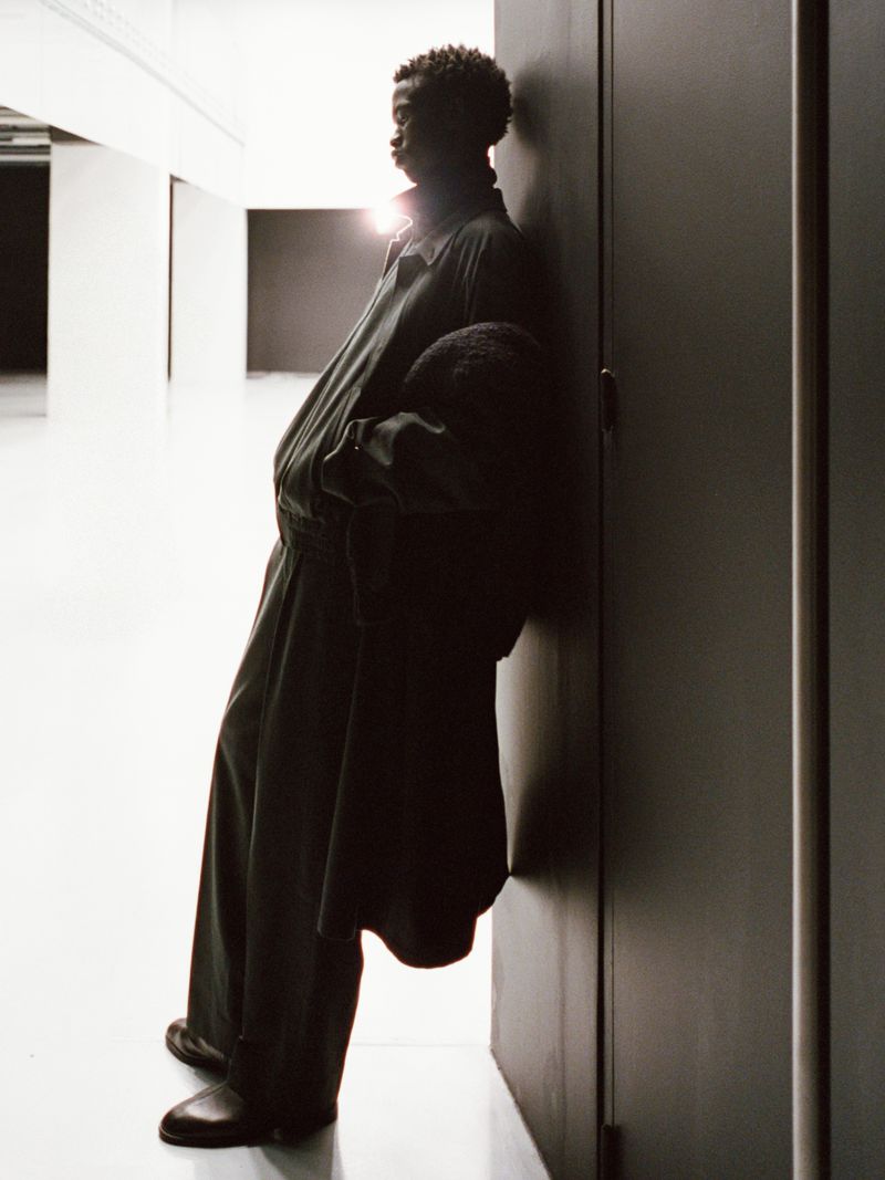 A slender African-descent adult leans against a dark door in loose fitting clothing inside a brightly illuminated hallway.