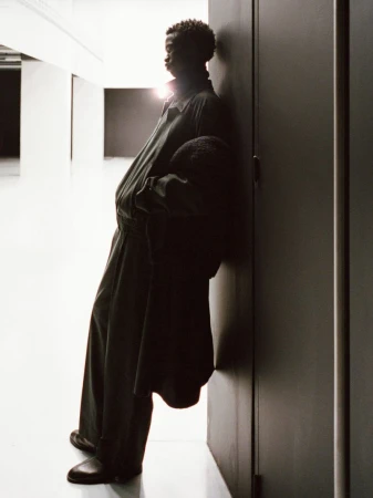A slender African-descent adult leans against a dark door in loose fitting clothing inside a brightly illuminated hallway.