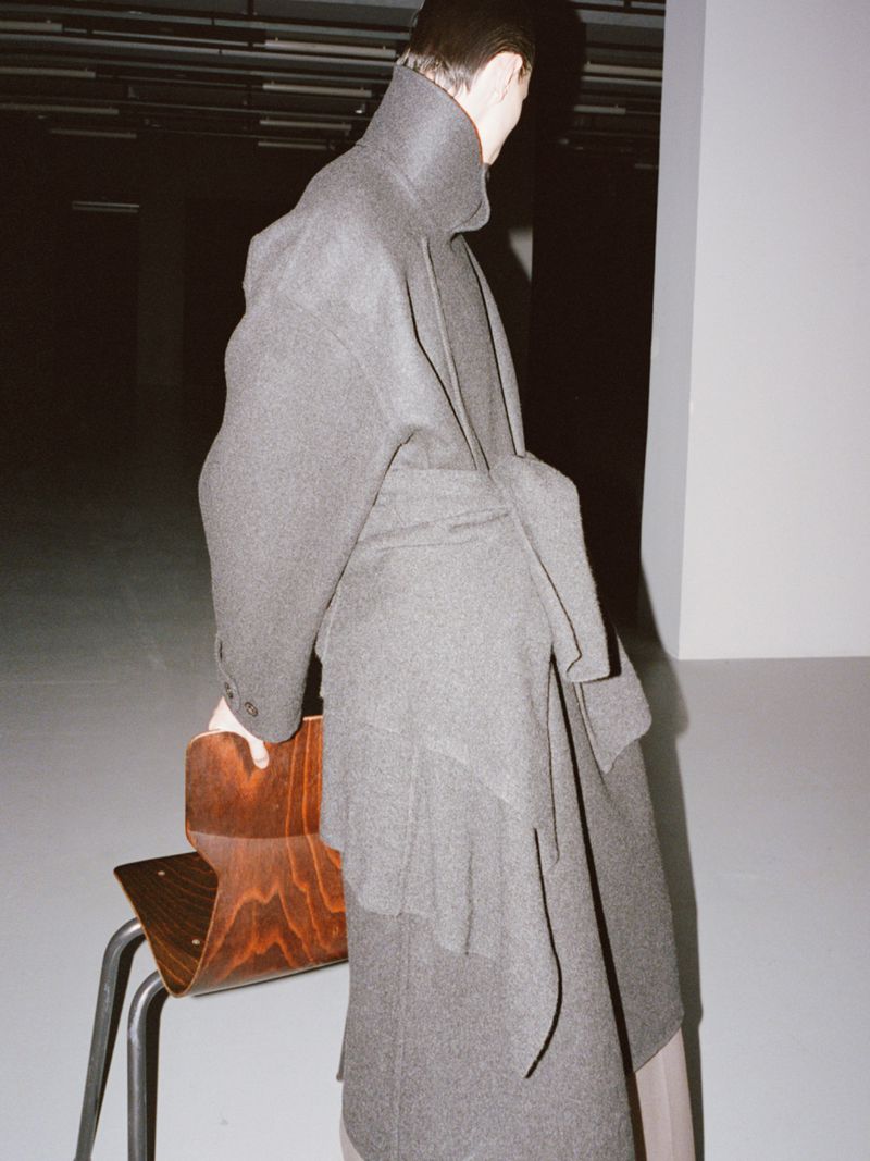 Stylish adult female wearing oversized grey coat with belt, posing indoors next to a wooden chair in a fashion or editorial shot.