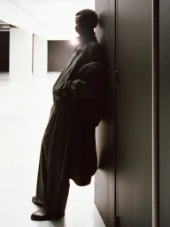 Model leaning against a dark partition wall wearing an oversized dark coat and wide-leg trousers in a minimalist space.