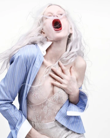 A female fashion model with silver hair in a studio setting poses in lingerie and an unbuttoned shirt with her mouth open and head tilted back.