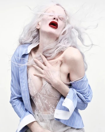 Fashionable woman with fair skin and gray hair poses with red lipstick wearing an open striped shirt and lace top in a studio setting.