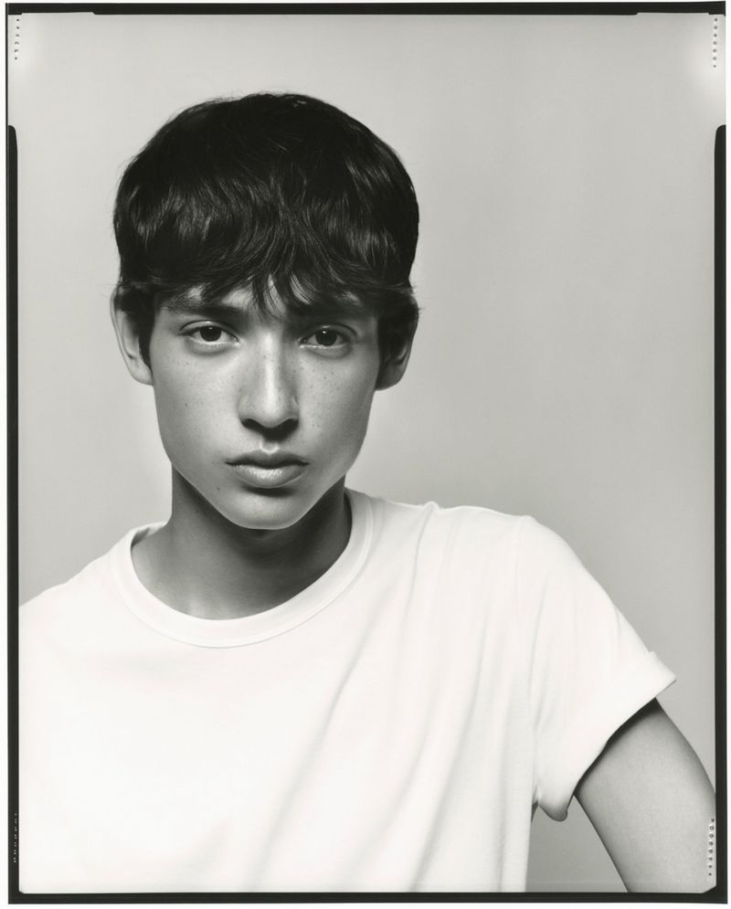 Portrait of a young adult with dark hair and light skin in a white t-shirt against a neutral background in a black and white photograph.