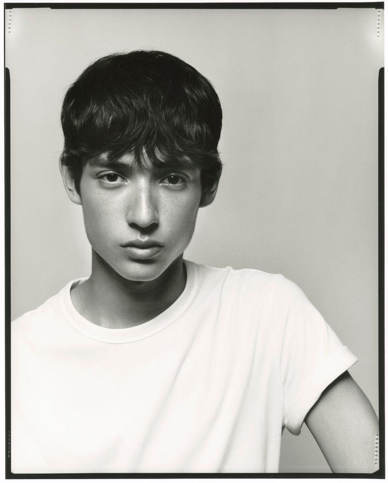 Monochrome portrait of a young adult with dark hair wearing a white T-shirt against a light grey backdrop in a photo studio.