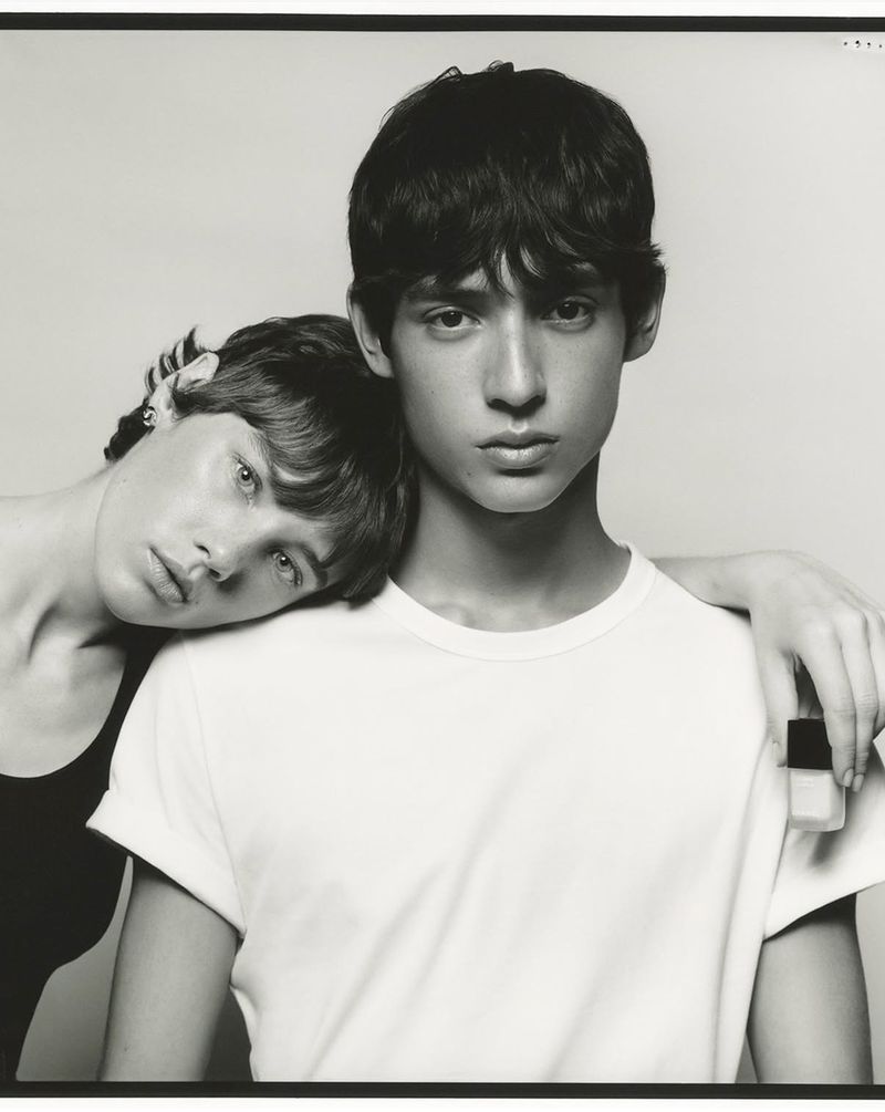 A black-and-white studio shot of a young couple: a female resting her head on the shoulder of a male wearing a white t-shirt.