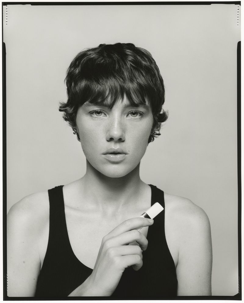 Monochrome portrait of a young adult female holding a white cosmetic tube against a neutral background in a studio setting.