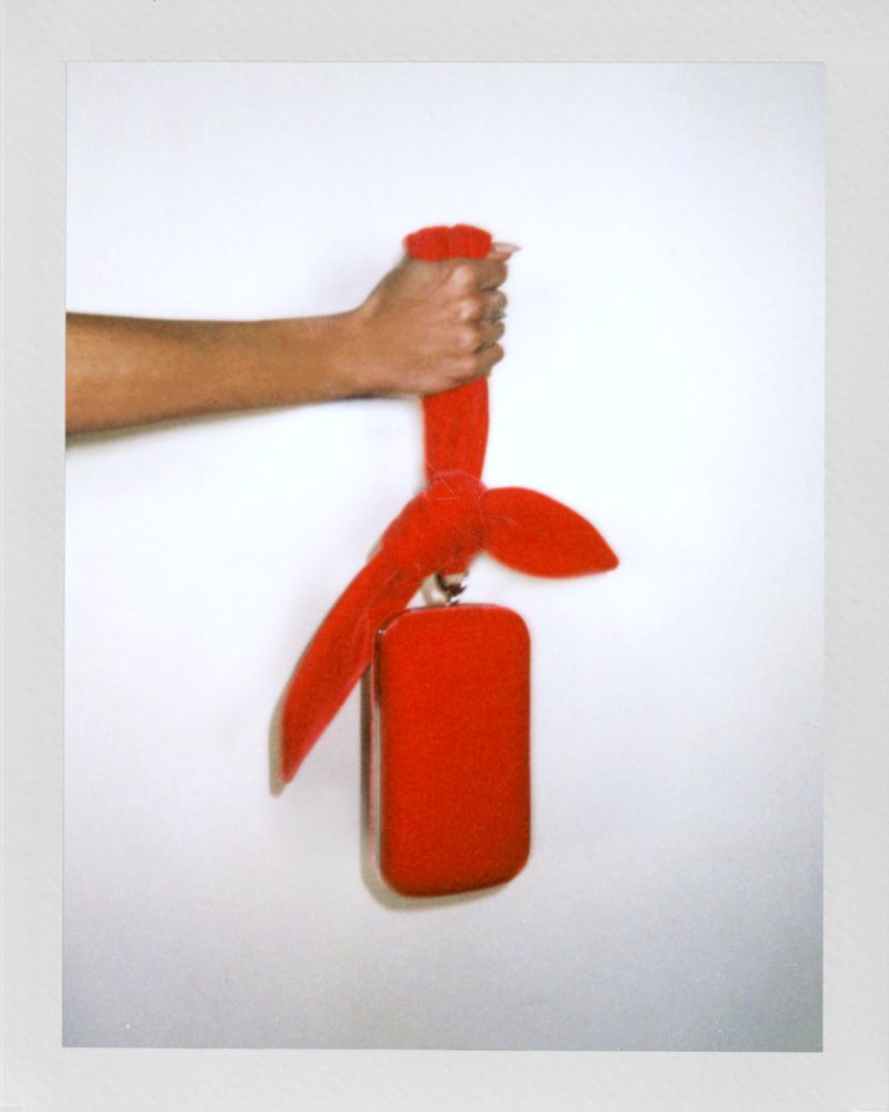 Arm holds up a rectangular red bag with red ruffled handle against a white background in a minimalist studio-lit fashion shot.