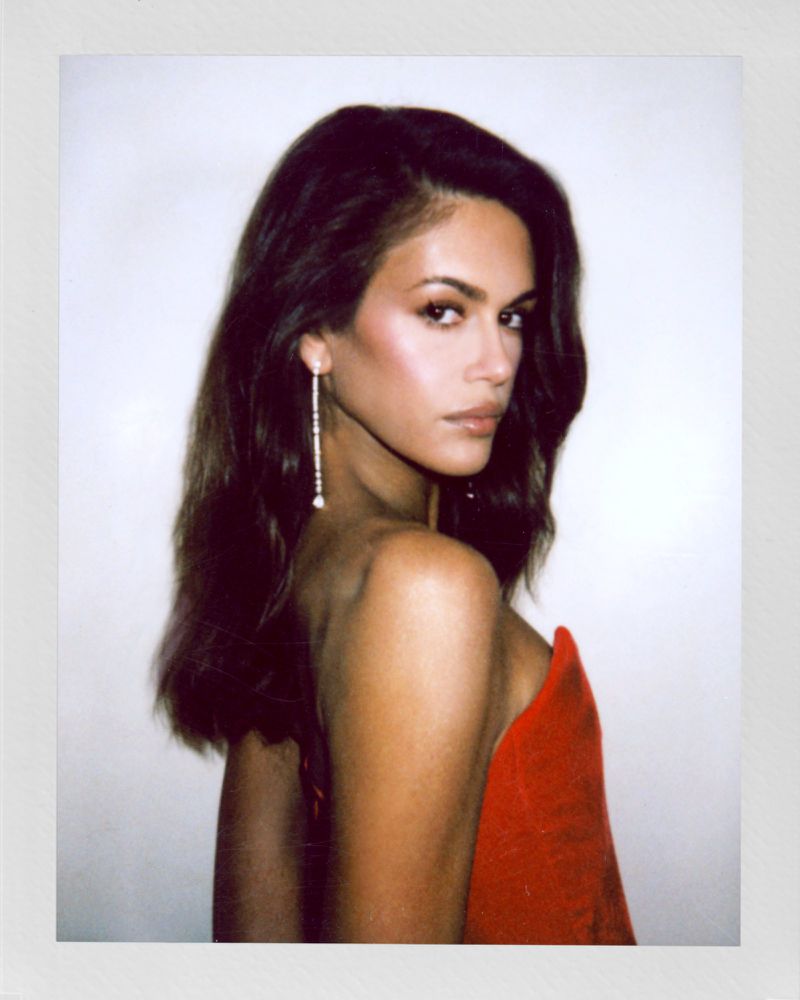 Close-up portrait of a woman with long brown hair, wearing red fabric and long earrings against a plain white backdrop.