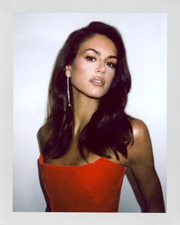Close-up of a dark-haired woman with dangling earrings in a red dress, posing against a white background in a studio setting.