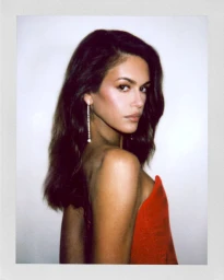 Model Kaia Gerber posing over her shoulder in a vibrant red strapless garment and crystal drop earring framed in a Polaroid.