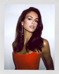 Polaroid portrait of Kaia Gerber in a red strapless top with a dangling earring against a white background.