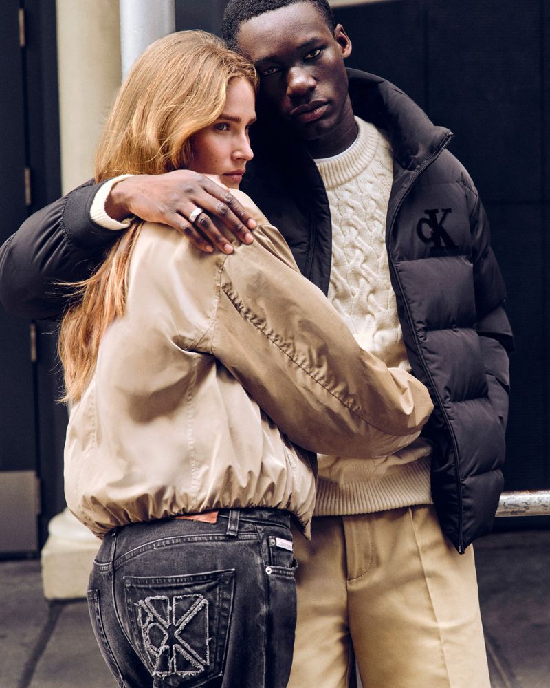 Fashion shot of a fair-skinned woman and a dark-skinned man in a close embrace, wearing jeans, puffer jackets, and neutral-toned pants and top.