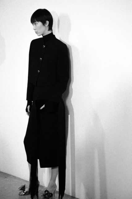 Monochromatic portrait of a stylish adult woman wearing a black jacket and skirt with fringe, and jeweled sandals against a white wall.