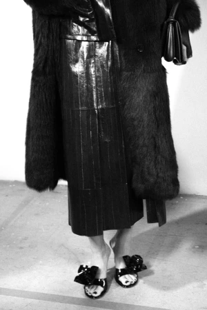 Model wearing a black fur coat, glossy midi skirt, and black bow heeled mules shown from the waist down in black and white.