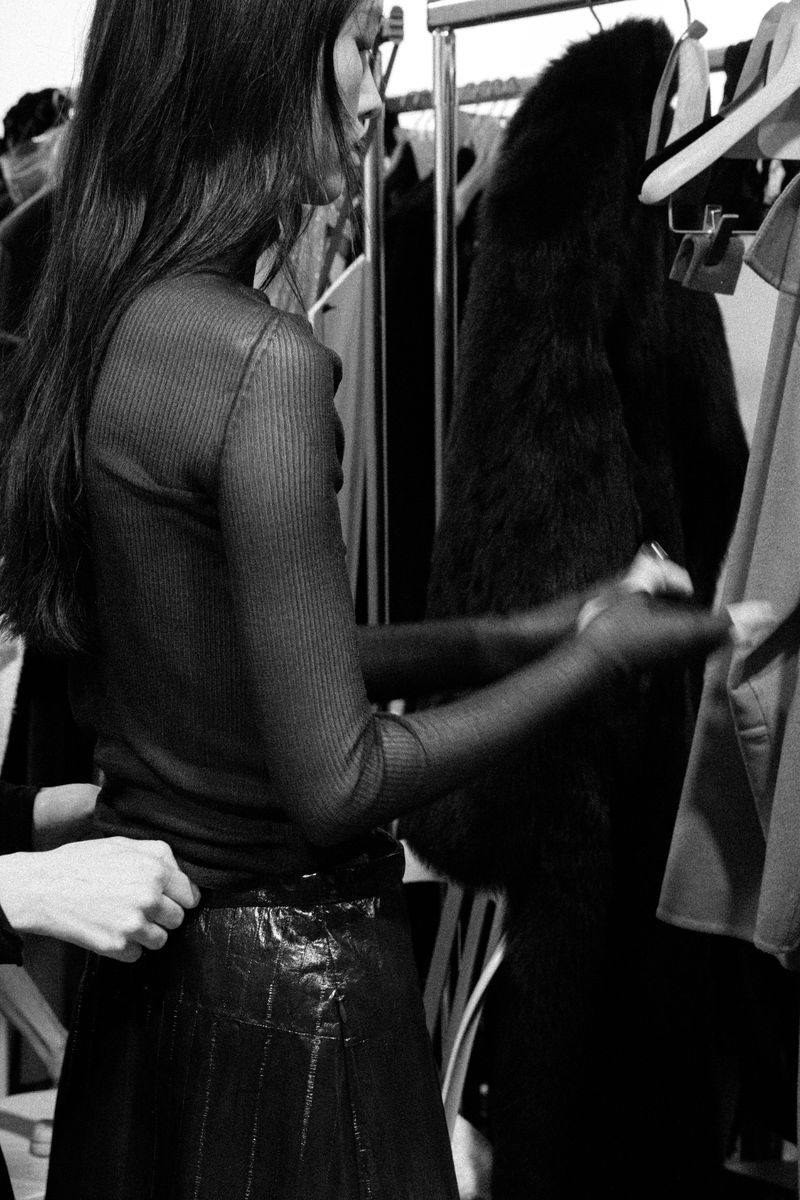 Black and white studio shot of a slender woman in a semi-sheer top and leather skirt, getting dressed in front of a rack of clothes.
