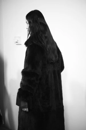 Model with long dark hair viewed from behind wearing an oversized black textured fur coat against a plain light wall.