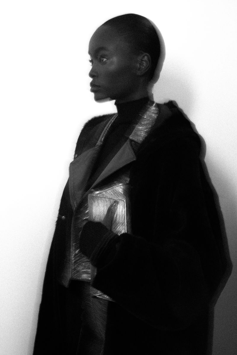 Monochrome portrait of a Black female model wearing a metallic jacket and black coat posing in profile against a white background.