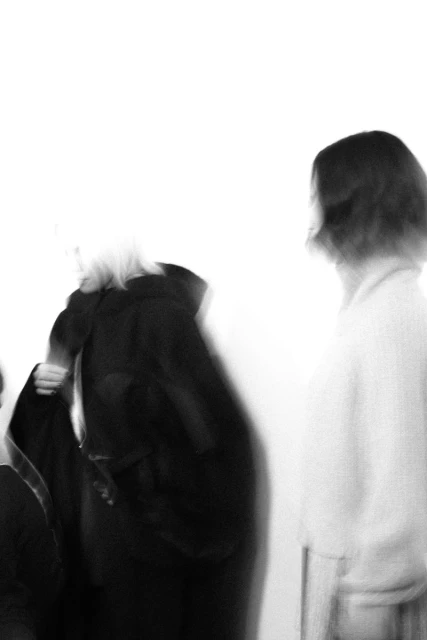 Two adult females in black and white tones; one figure wearing a long black coat in soft focus against bright white background.