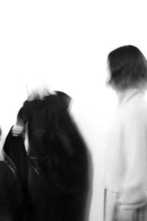Two adult females in black and white tones; one figure wearing a long black coat in soft focus against bright white background.
