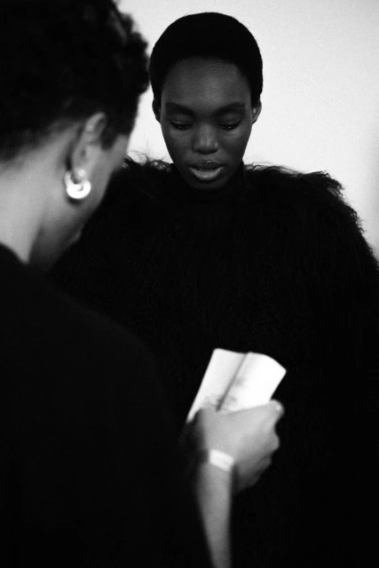 Black and white fashion portrait of two African-descent women, one holding a pamphlet or brochure, in a minimalist studio setting.
