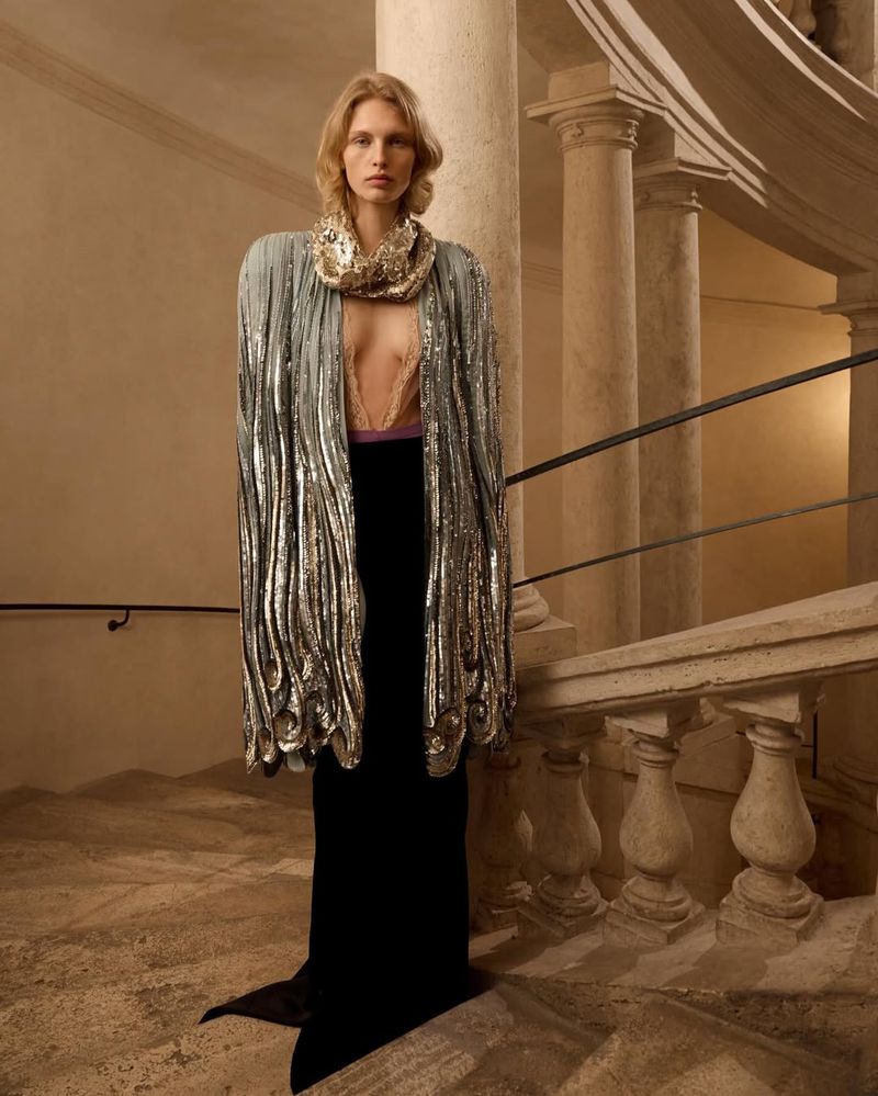 Model wearing a shimmering silver sequined cape and black skirt standing on an ornate stone staircase with classical columns.