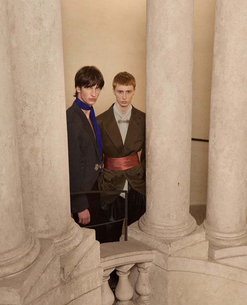 Two young male fashion models pose between marble columns in suits with ascots and cummerbunds against a neutral background.