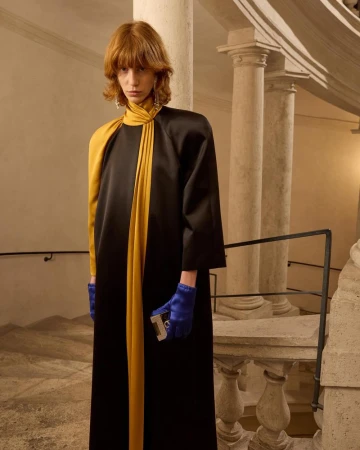 Fashionable red-haired model wearing long black and gold dress with blue gloves posing on a staircase with stone pillars inside.