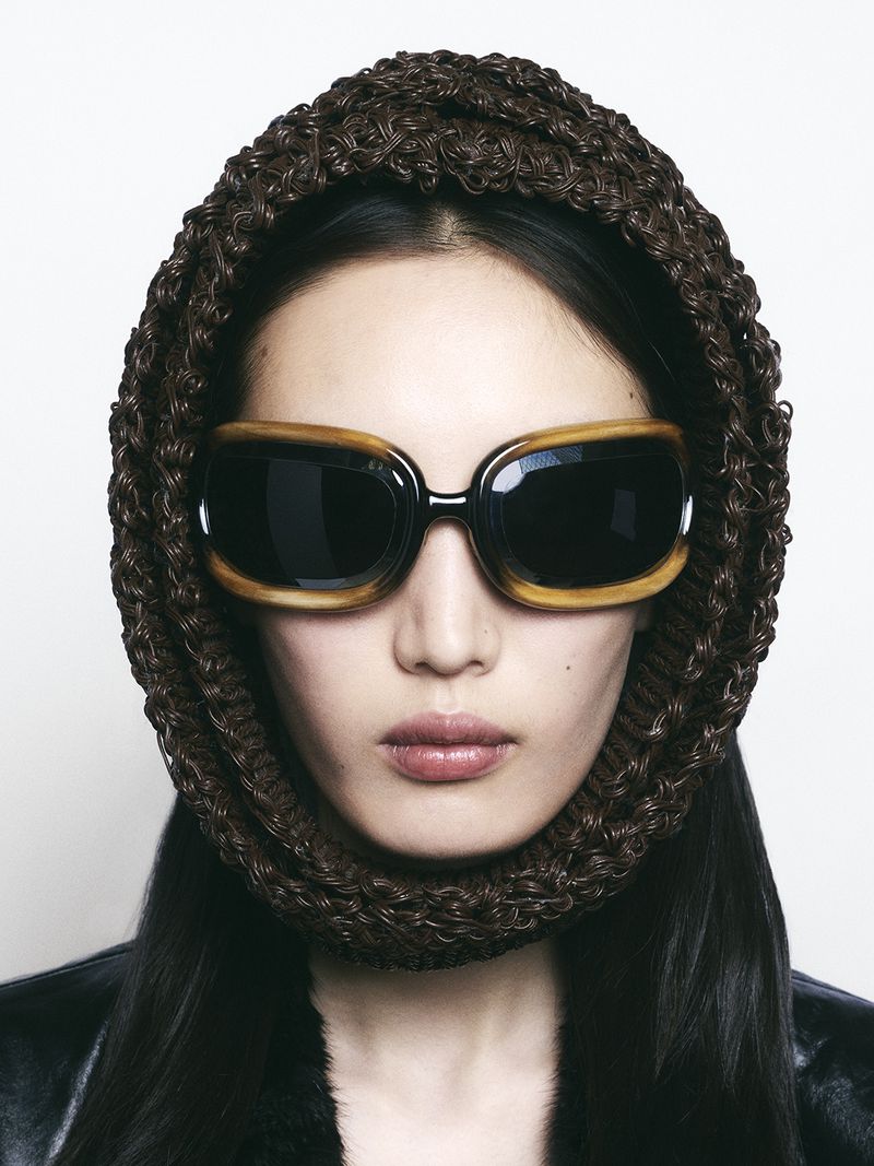 Portrait of a woman wearing sunglasses and a brown woven head covering. She is wearing a black leather jacket and light makeup.