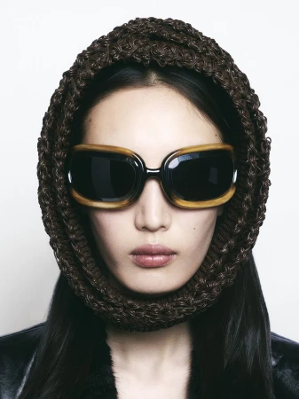 Portrait of a woman wearing sunglasses and a brown woven head covering. She is wearing a black leather jacket and light makeup.