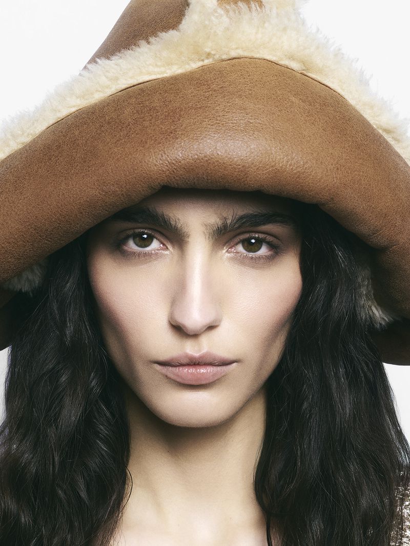Close-up portrait of a woman wearing a brown fur-trimmed hat against a clean white background in a studio setting, beauty shot.