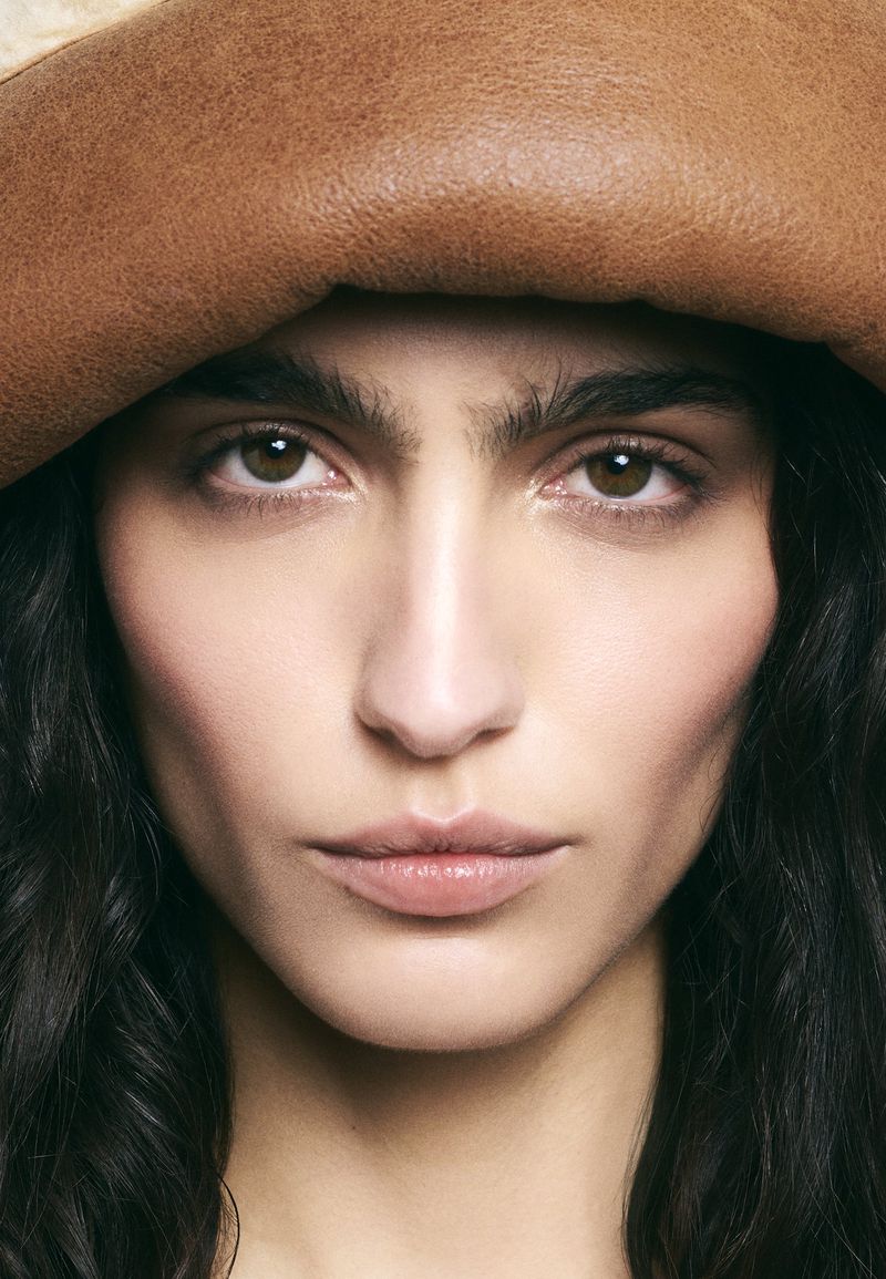 Close-up portrait of a woman with brown eyes and dark hair wearing a brown hat-like object looking directly at the camera.