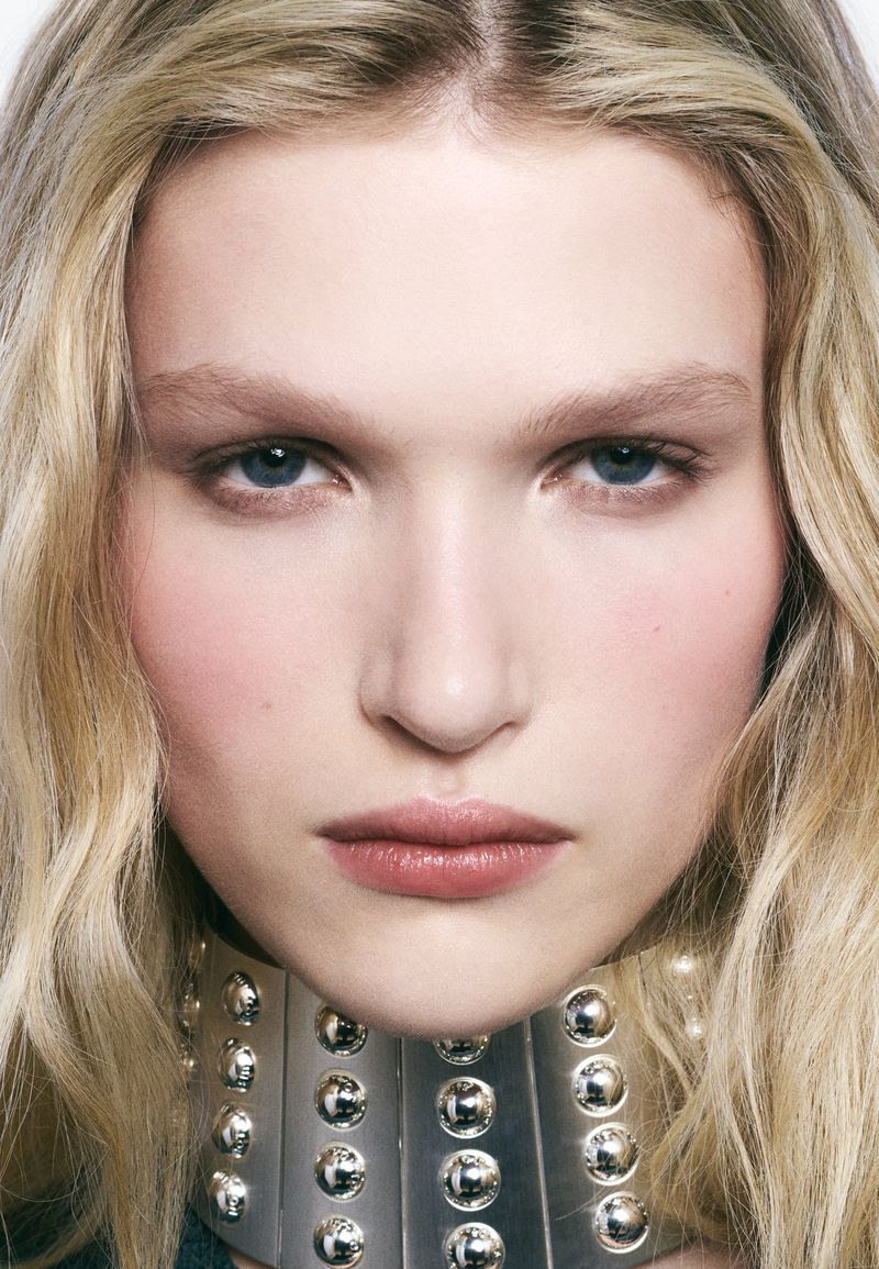 Close-up portrait of a young fair-skinned female model with wavy blonde hair wearing silver studded collar necklace in a photo studio.