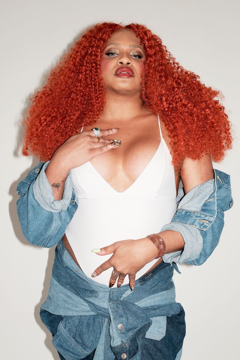 A person with voluminous vibrant red curly hair wears a white bodysuit, denim jacket, and denim skirt against a plain light background.