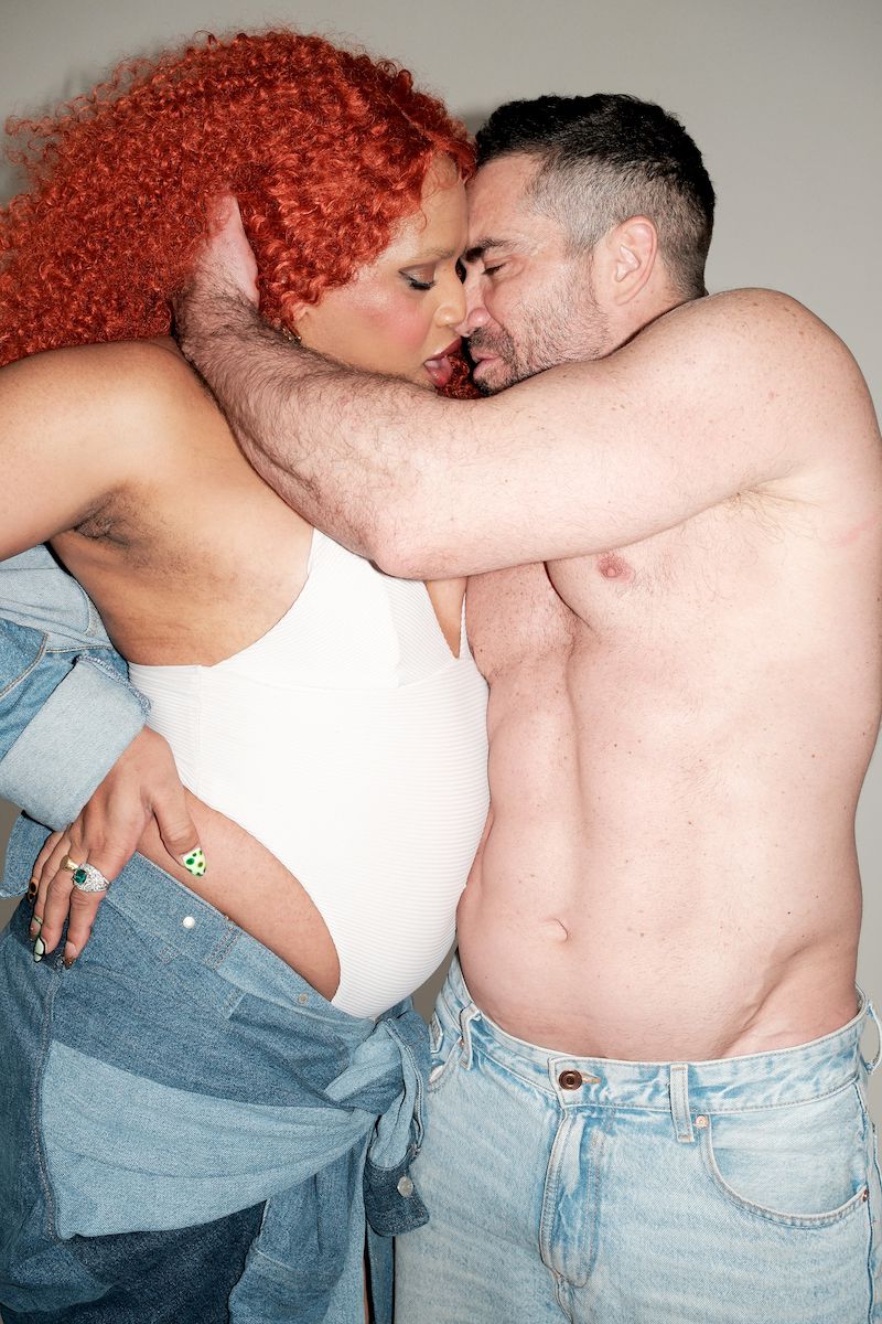 A couple embraces in a close-up studio shot. The woman has red curly hair and wears a white bodysuit and denim, while the man is shirtless.