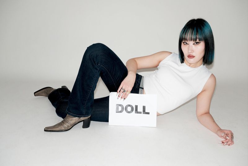 A woman with blue-streaked hair poses while holding a sign that reads 'DOLL'. She wears a white top, jeans, and heeled boots.