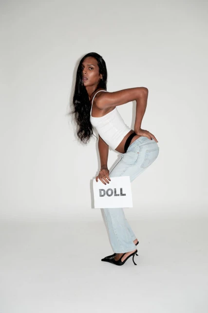 Transgender woman in casual outfit poses in a photo studio holding a sign that reads 'DOLL' in black block letters.