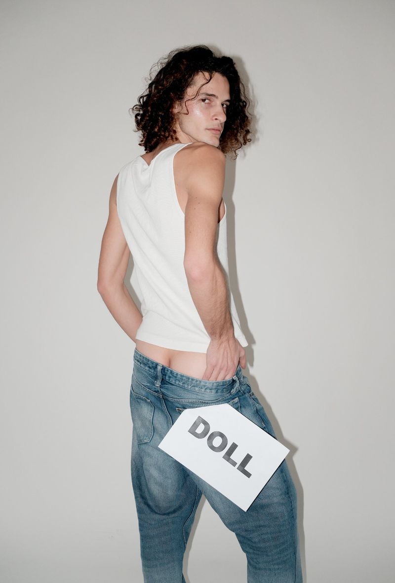 Man in jeans poses in a photo studio holding a sign that says DOLL in a fashion shot. He is looking over his shoulder.