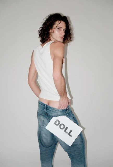 Man in jeans poses in a photo studio holding a sign that says DOLL in a fashion shot. He is looking over his shoulder.