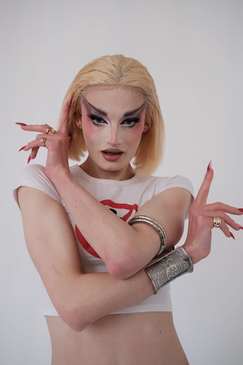 Striking close-up portrait of a blonde adult wearing elaborate drag makeup, a white cropped t-shirt, arm cuffs, and rings against a white background.