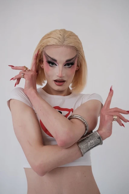 Striking close-up portrait of a blonde adult wearing elaborate drag makeup, a white cropped t-shirt, arm cuffs, and rings against a white background.