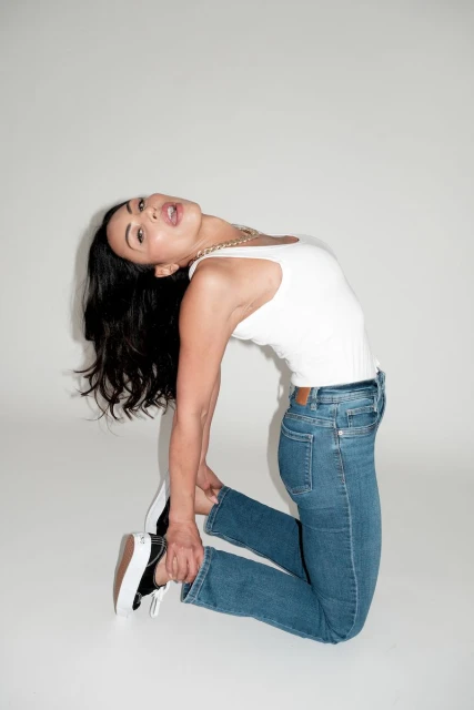 A woman with dark hair arches backwards in a white ribbed tank top and blue jeans, wearing black and white sneakers against a white backdrop.