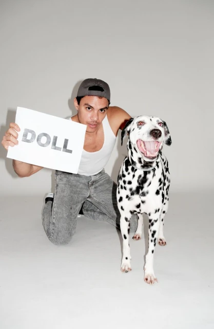 Dalmatian dog and a man holding a sign that says 'Doll' in a studio. The man is kneeling and wearing a baseball cap and a tank top.