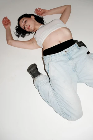 Woman with dark curly hair lying on a white background wearing a white crop top and light blue denim jeans.