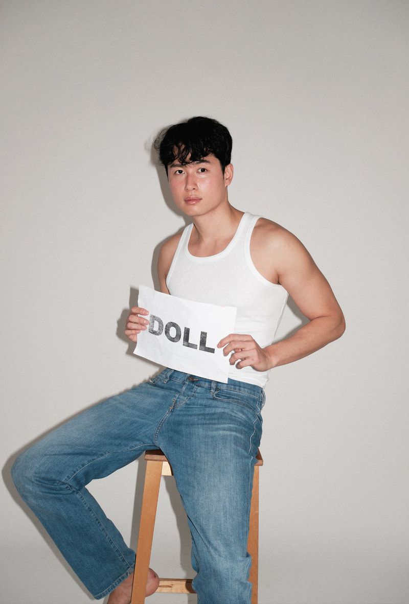 An adult male seated on a wooden stool in a studio setting, wearing a white tank top and blue jeans while holding a sign that reads 'DOLL'.