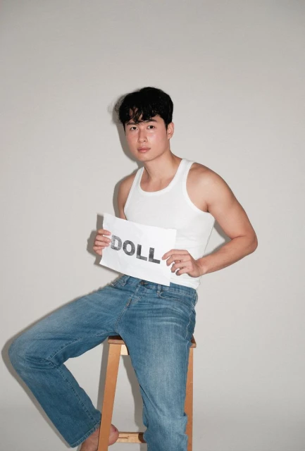 An adult male seated on a wooden stool in a studio setting, wearing a white tank top and blue jeans while holding a sign that reads 'DOLL'.