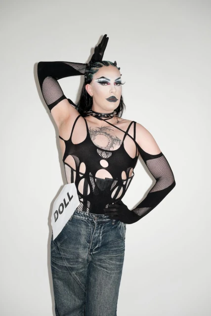 Drag queen with makeup poses in a black outfit and denim jeans with white background in studio lighting.