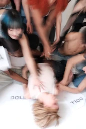 Top-down view of fashion models with dynamic motion blur surrounding a person lying on the floor next to paper signs reading DOLL.