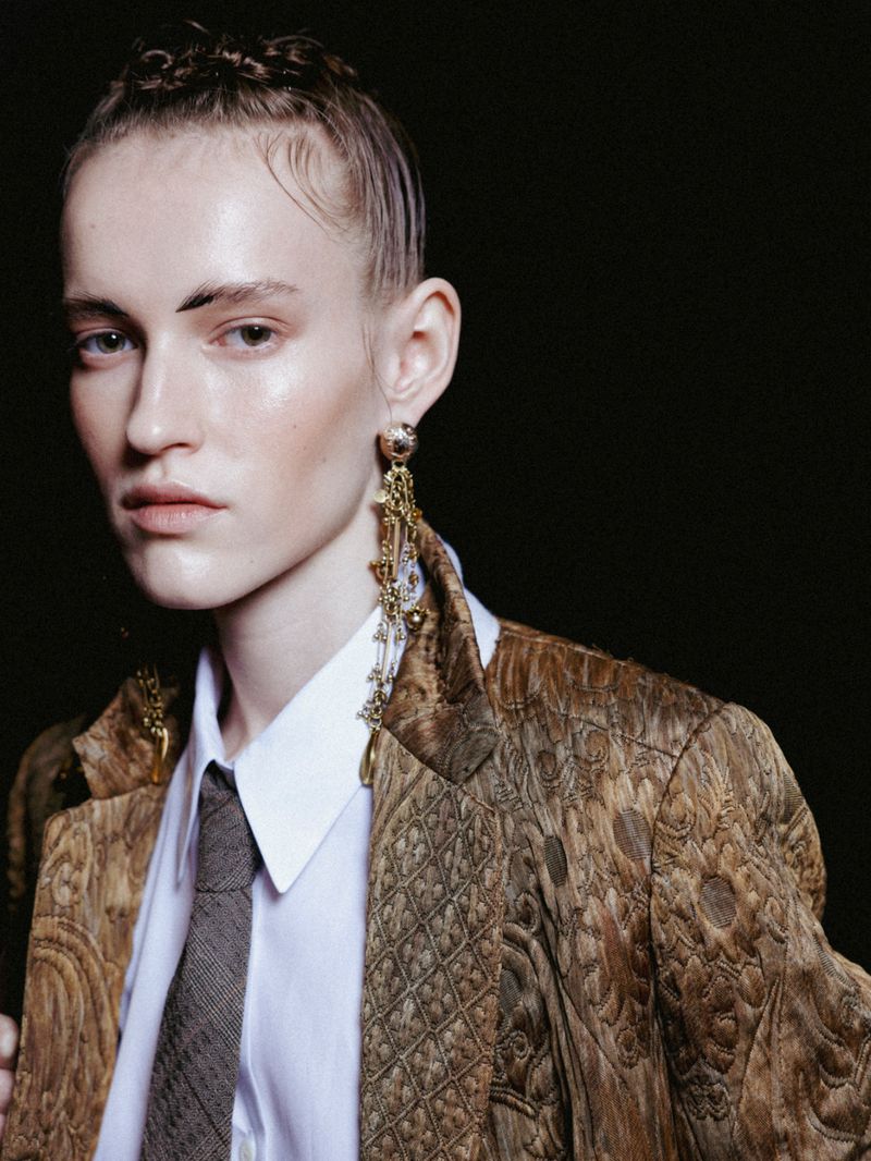Portrait of adult female with patterned blazer, collared shirt, and tie against a dark background in a fashion editorial style.
