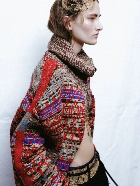 Profile view of a fashion model wearing a patterned brown, red, and blue sweater, ornate jewelry, and a braided hairstyle in a studio setting.