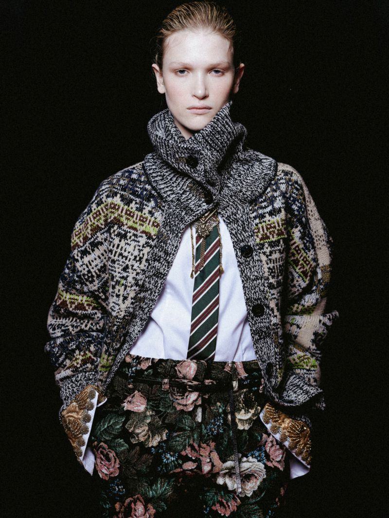 Fashion editorial shot of a young adult woman in a patterned coat and floral pants against a stark black backdrop in a studio setting.